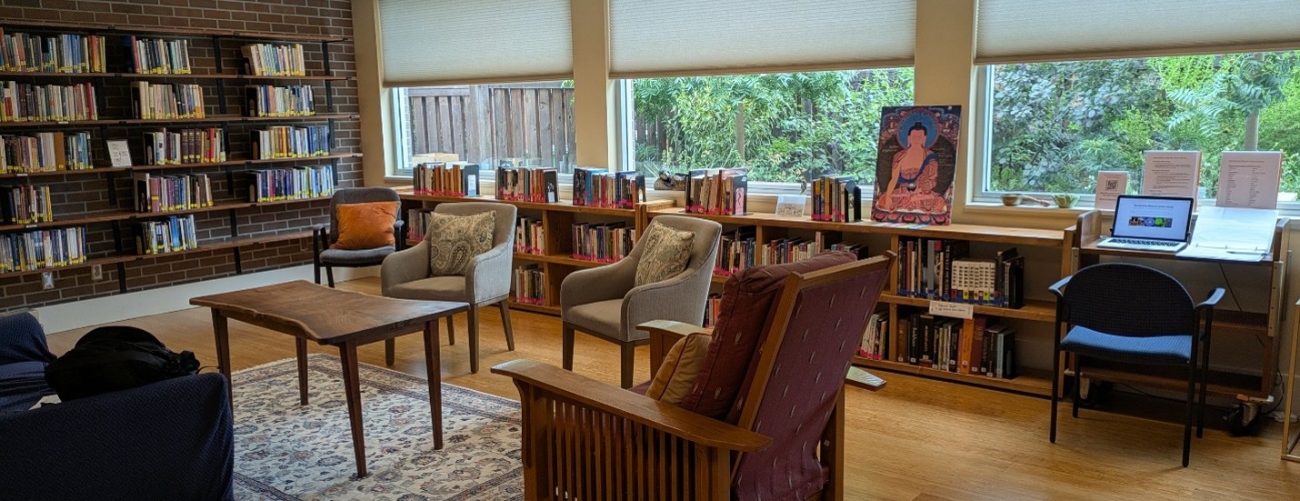 a photograph of the inside of the Sacramento Dharma Center Library showing bookshelves, windows, chairs, a small table, a laptop, and a painting of Buddha in the earth touching gesture.